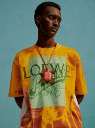 Portrait of a Black man wearing a yellow graphic t-shirt and a crab pendant necklace against a blue textured background.