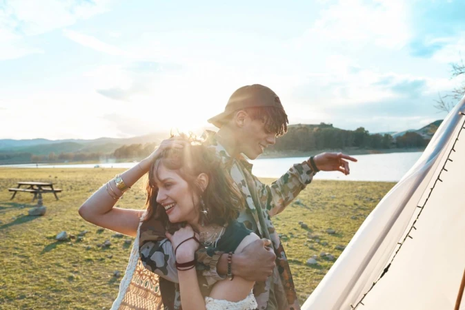 Couple embraces near tent on grassy field with lake and hills. Woman smiles, man points. Bright sunny day, outdoor fashion.
