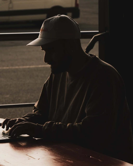 Silhouetted view of an adult man typing on a laptop at an outdoor table with a baseball cap, sweatshirt, and natural light.