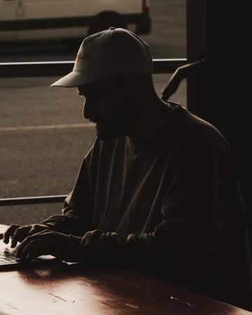 Silhouetted view of an adult man typing on a laptop at an outdoor table with a baseball cap, sweatshirt, and natural light.