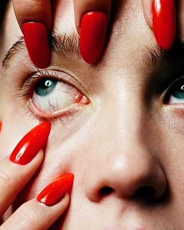 Close-up of a woman's face with striking blue eyes and red manicured fingernails touching her face in a beauty portrait.