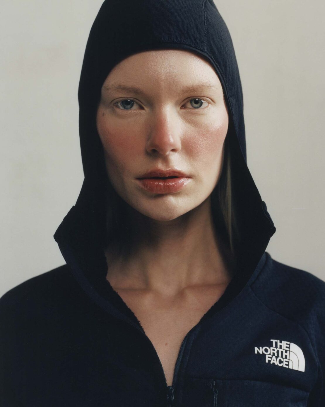 Close-up studio shot of a fair-skinned woman wearing a black hood and a black and navy blue zip-up jacket, against a neutral background.