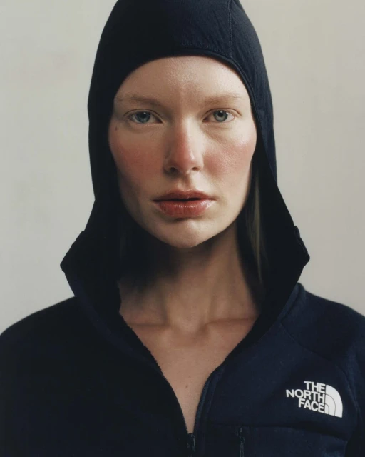 Close-up studio shot of a fair-skinned woman wearing a black hood and a black and navy blue zip-up jacket, against a neutral background.