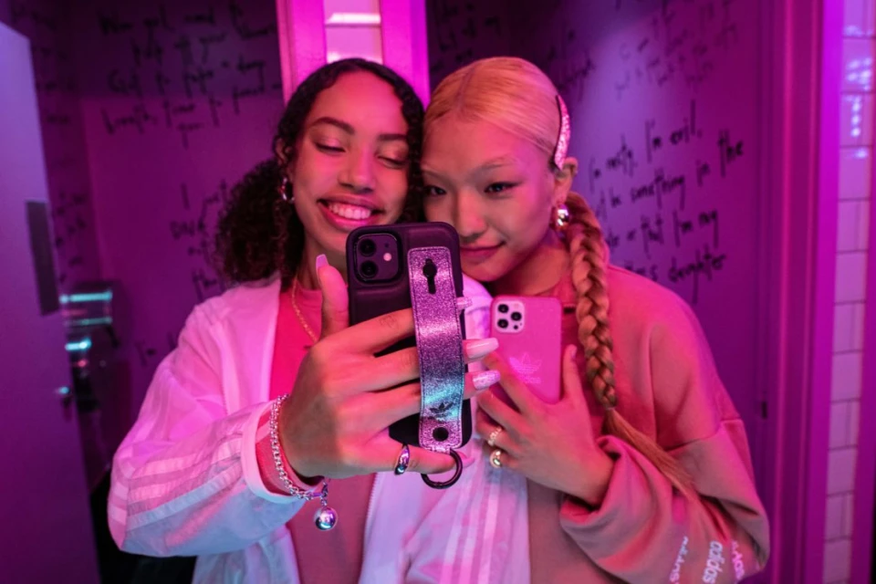 Two smiling young women with phones pose indoors under purple lighting. One woman wears Adidas branded apparel.