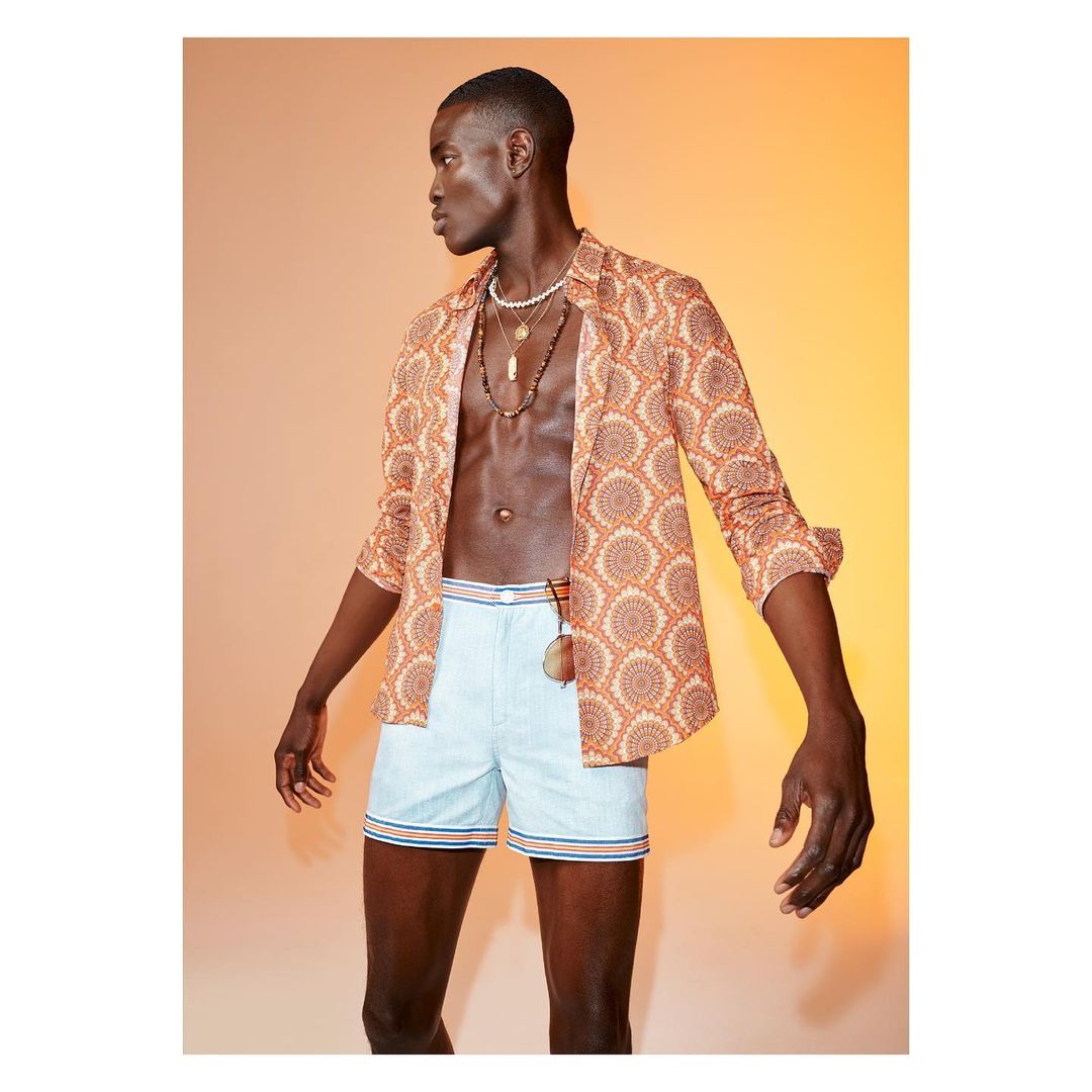 Male model posing in patterned shirt and striped shorts, accessorized with layered necklaces against a gradient background.