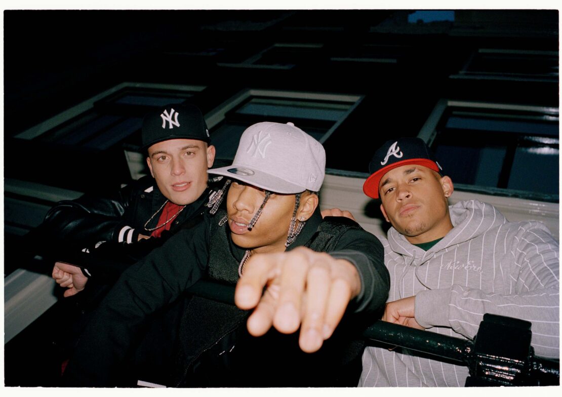 Three stylish young men pose in baseball caps, hoodies, and jackets against a windowed building at night, showcasing urban fashion.