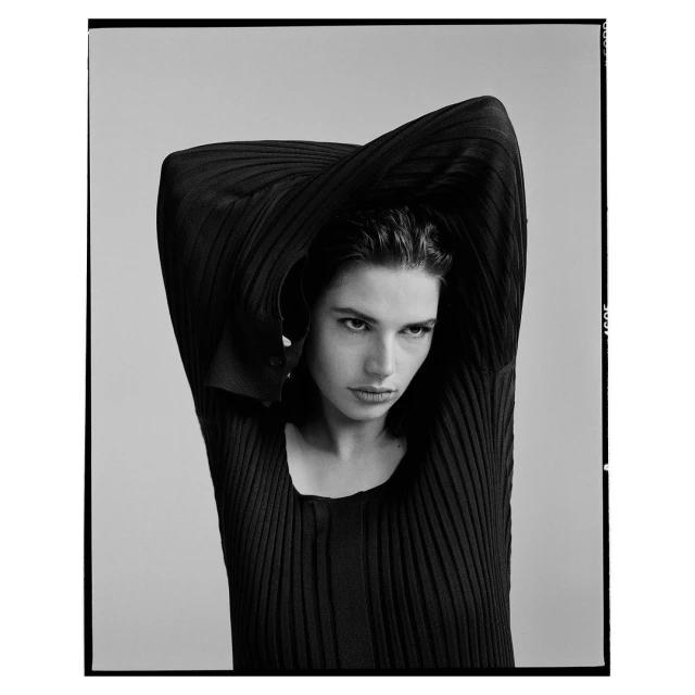 Monochrome portrait of a brunette woman in a ribbed top with arms raised, set against a neutral backdrop in a studio.