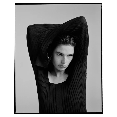 Monochrome portrait of a brunette woman in a ribbed top with arms raised, set against a neutral backdrop in a studio.