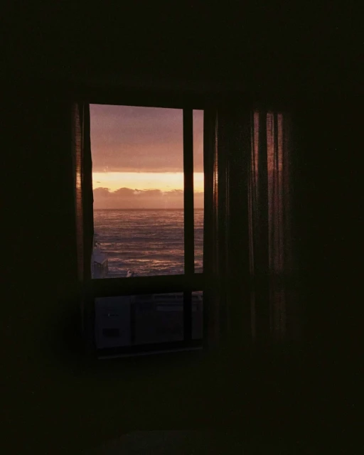 View of the ocean at dusk through a window with drawn curtains and a visible air conditioner, creating a moody, indoor scene.