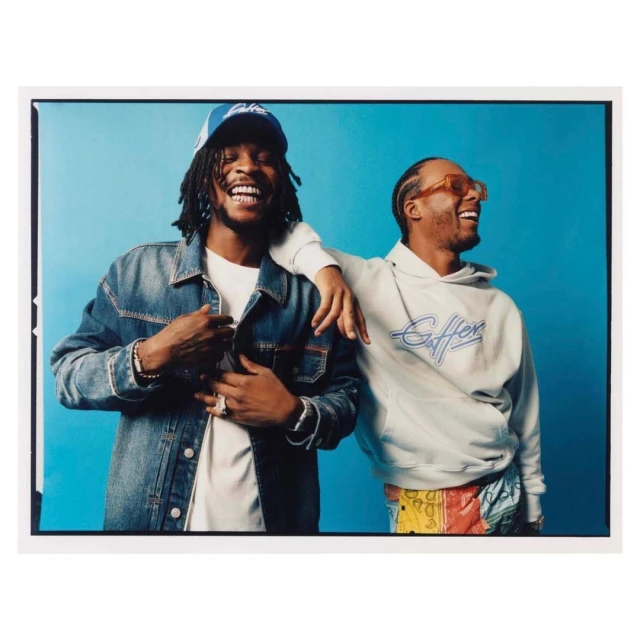 Two smiling adult males pose against a blue backdrop, showcasing a denim jacket, hoodie, baseball cap, and stylish accessories, indoor studio.
