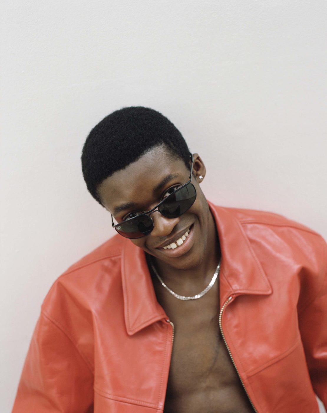 Portrait of an adult Black male smiling and wearing sunglasses and a red leather jacket against a pale background in a medium shot.