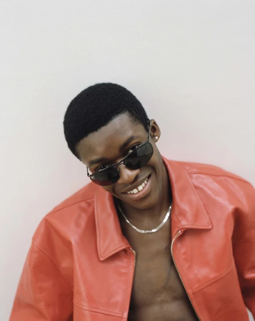 Portrait of an adult Black male smiling and wearing sunglasses and a red leather jacket against a pale background in a medium shot.