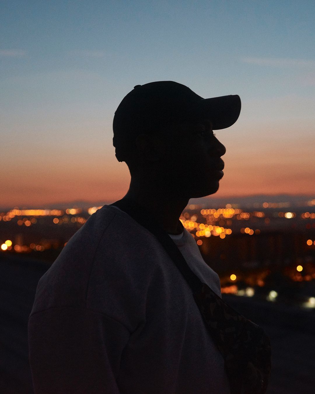 Silhouette of an adult male wearing a cap and crossbody bag against a twilight sky with a blurred cityscape in the distance.