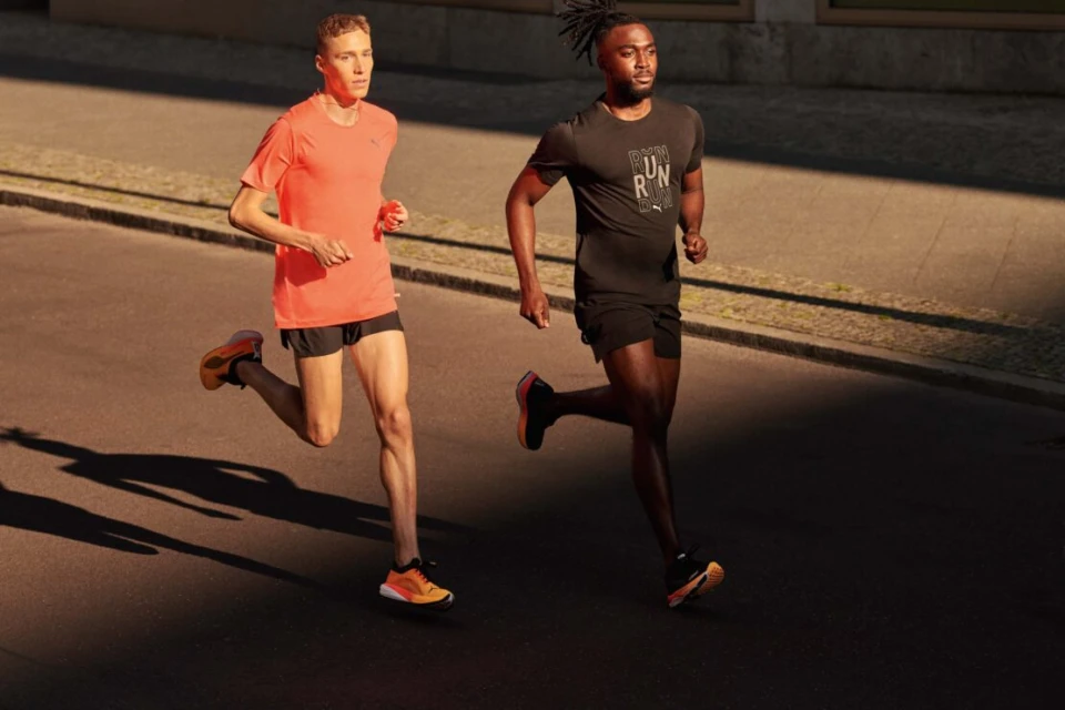 Two athletic men run on a sunny city street. One man wears an orange tee, the other a black Puma tee and black shorts, athletic shoes.
