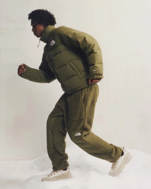 An adult male model runs in an olive green puffer jacket and fleece pants on a white set, promoting active fashion and outdoor apparel.