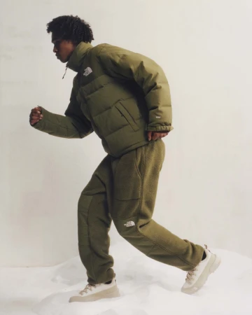 An adult male model runs in an olive green puffer jacket and fleece pants on a white set, promoting active fashion and outdoor apparel.