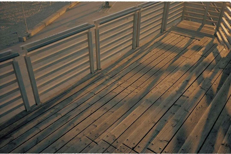 Elevated wooden walkway with railings casting shadows, blending architectural design with natural light and surface texture.