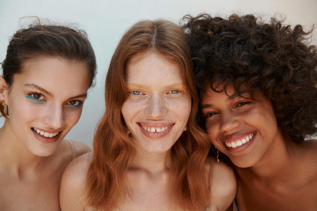 Close-up portrait of three smiling young women with distinctive hair and makeup against a pale background by Elliott Wilcox.