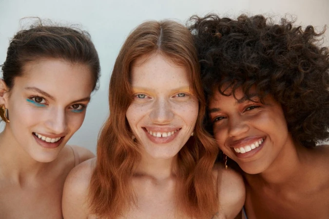 Close-up portrait of three smiling young women with distinctive hair and makeup against a pale background by Elliott Wilcox.