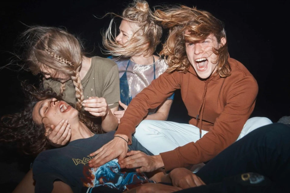 A group of four young adults are captured in a candid moment with one person's hand touching another person's face, against a dark background.
