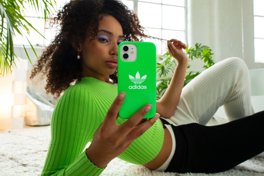 Young woman poses indoors with a green Adidas-branded cell phone case, wearing a green sweater and white pants, in a lifestyle shot.
