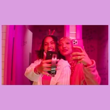 Two young women taking a selfie with pink smartphones, posing indoors with pink and purple lighting, smiling and making gestures.