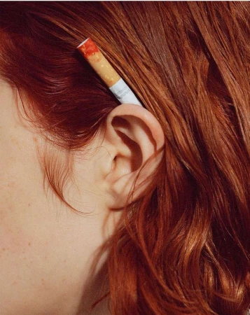 Close-up portrait of a person with red hair tucked behind their ear, holding a cigarette
