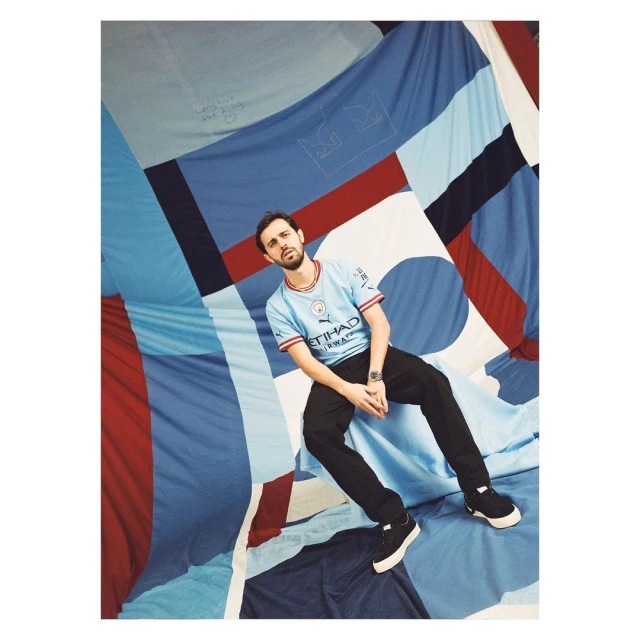 Adult male in a sky-blue jersey sits against a geometrically patterned backdrop, posing in black pants and sneakers for a portrait.
