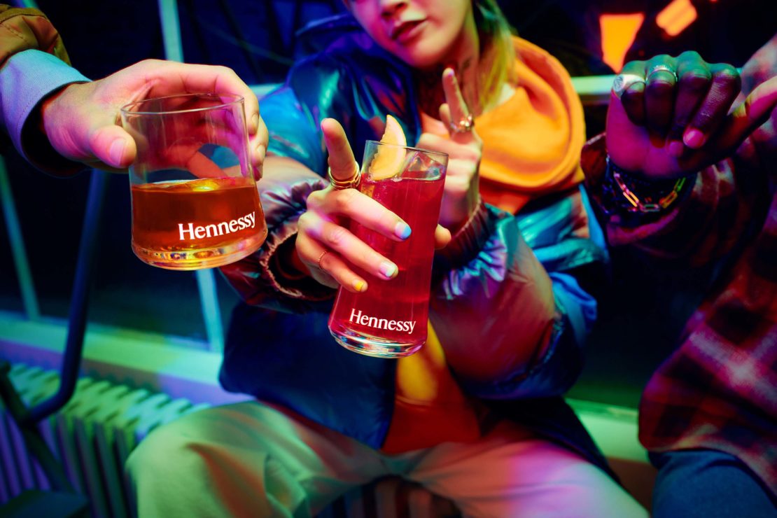 Three people hold up cocktail glasses with Hennessy branding. Indoor setting with blue and green neon lighting creates a vibrant mood.