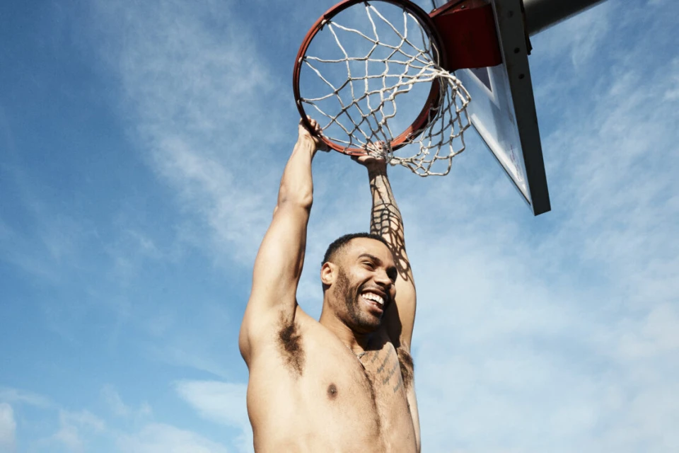 Athletic man hanging from a basketball hoop against a blue sky. He is smiling, shirtless, and has tattoos on his arm.