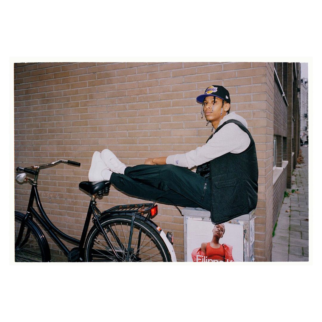 Young adult male in baseball cap and vest poses seated on a bike in an urban alleyway against a brick wall near a poster.
