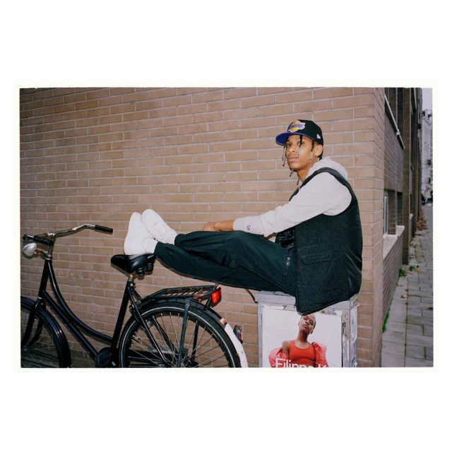 Young adult male in baseball cap and vest poses seated on a bike in an urban alleyway against a brick wall near a poster.