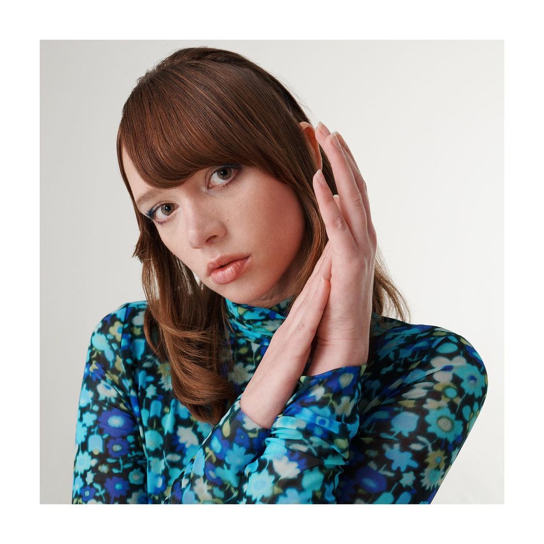 Portrait of a young adult woman with brown hair, wearing a floral patterned top, posing with her hands near her face against a white backdrop.