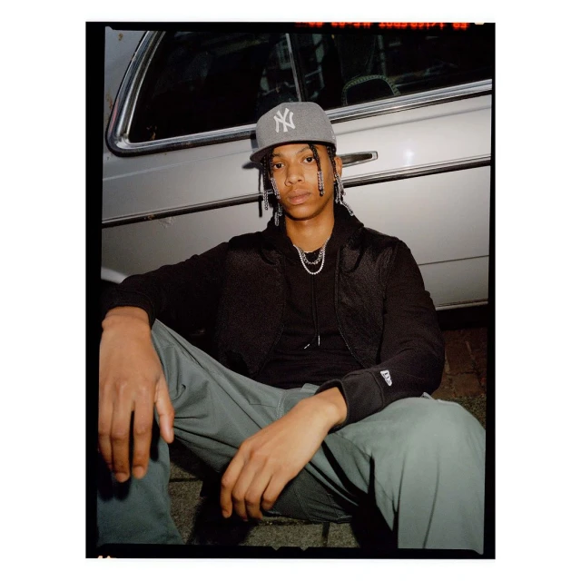 Portrait of a young man wearing a baseball cap and jewelry, sitting outdoors with a silver car partially visible in the background.