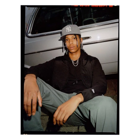 Portrait of a young man wearing a baseball cap and jewelry, sitting outdoors with a silver car partially visible in the background.