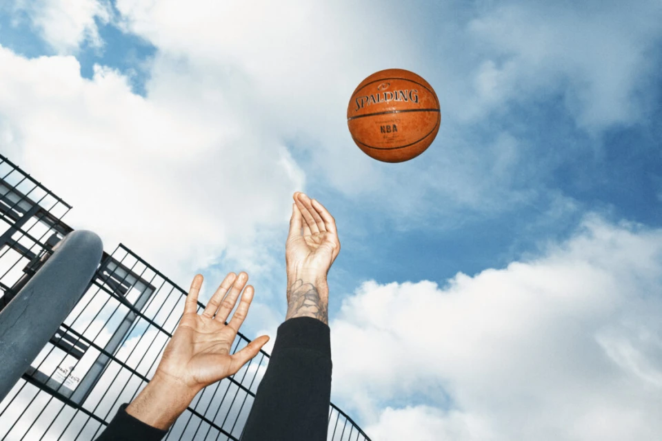 Low angle view of an orange Spalding NBA basketball floating in the air as an arm reaches up, set against a background of sky and building.