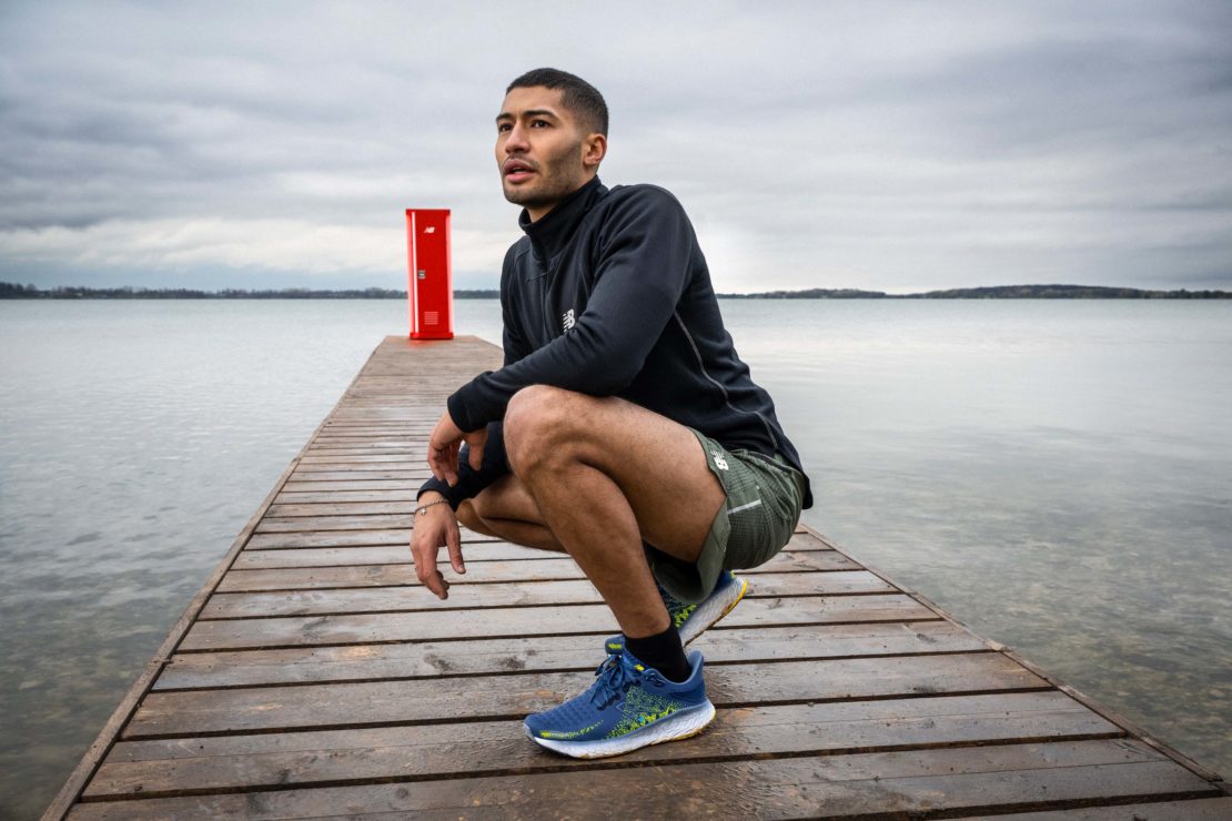 Athletic man in workout gear squatting on a wooden pier with a red post, against a cloudy sky and a tranquil body of water.