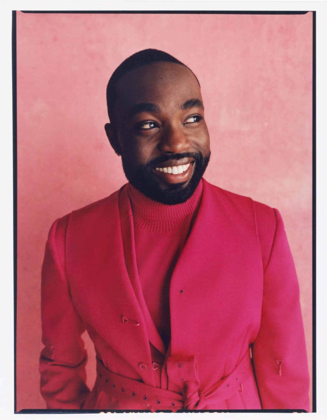 Smiling African-American man in a pink suit and turtleneck sweater looking off-camera in a portrait on a pink background.