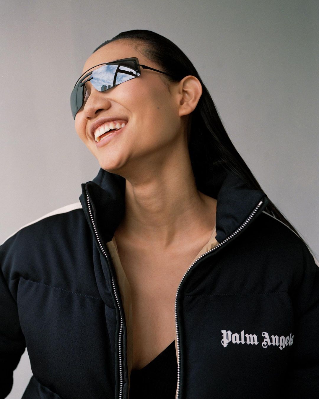 Smiling woman wearing sunglasses and a black Palm Angels puffer jacket poses against a light-grey background in a close portrait.