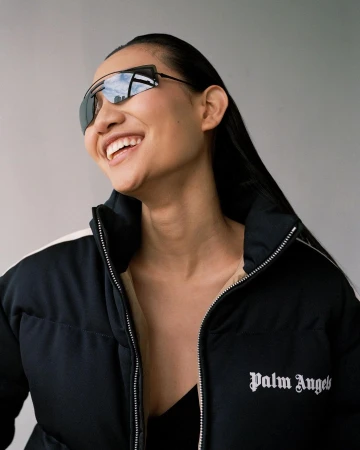 Smiling woman wearing sunglasses and a black Palm Angels puffer jacket poses against a light-grey background in a close portrait.
