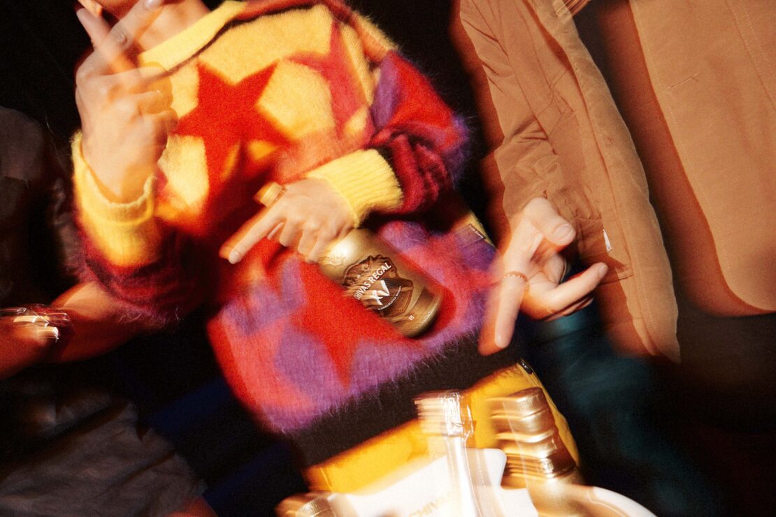 Close-up of two people at a party, one wearing a multicolored star sweater and holding a champagne bottle in a blurry, energetic photo.