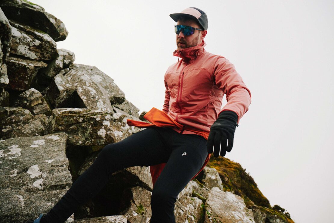 Man trail running on a rocky mountain in sportswear. He wears a light red jacket, cap, sunglasses, and gloves for outdoor fitness.