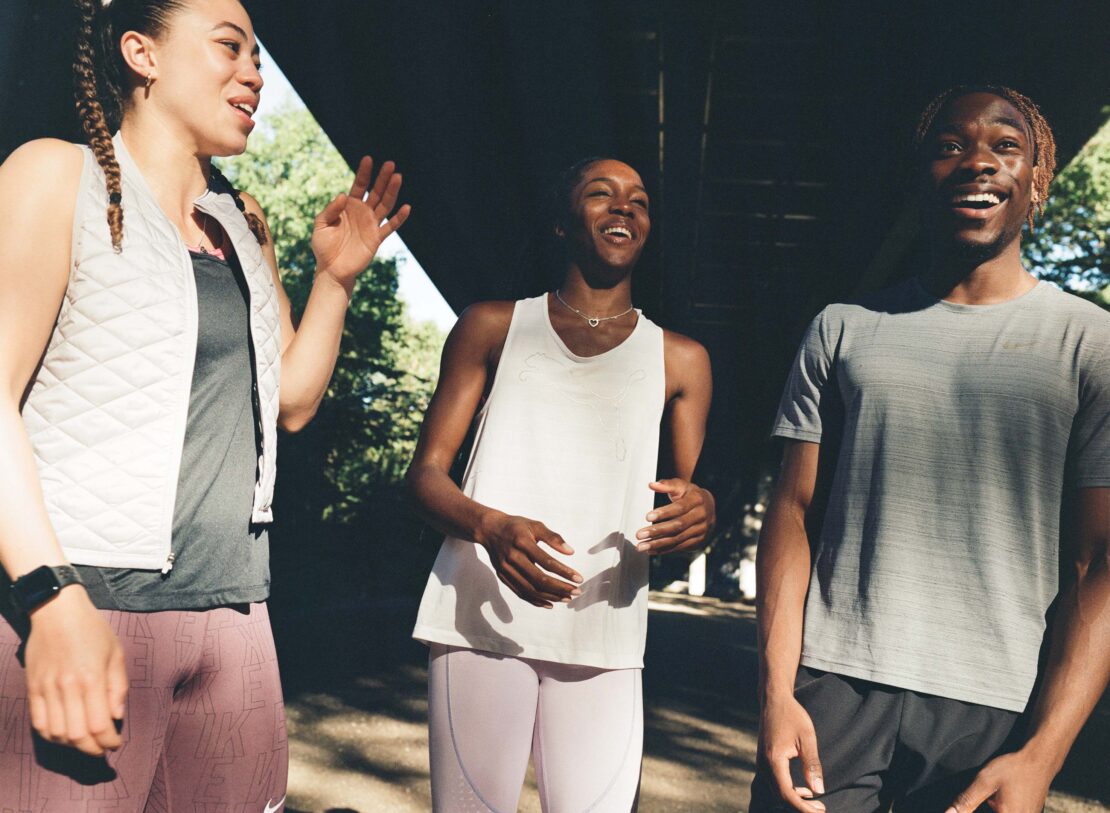 Three smiling athletes, two women and one man, wear athletic wear in an outdoor environment with dark shadows and sunlight. 