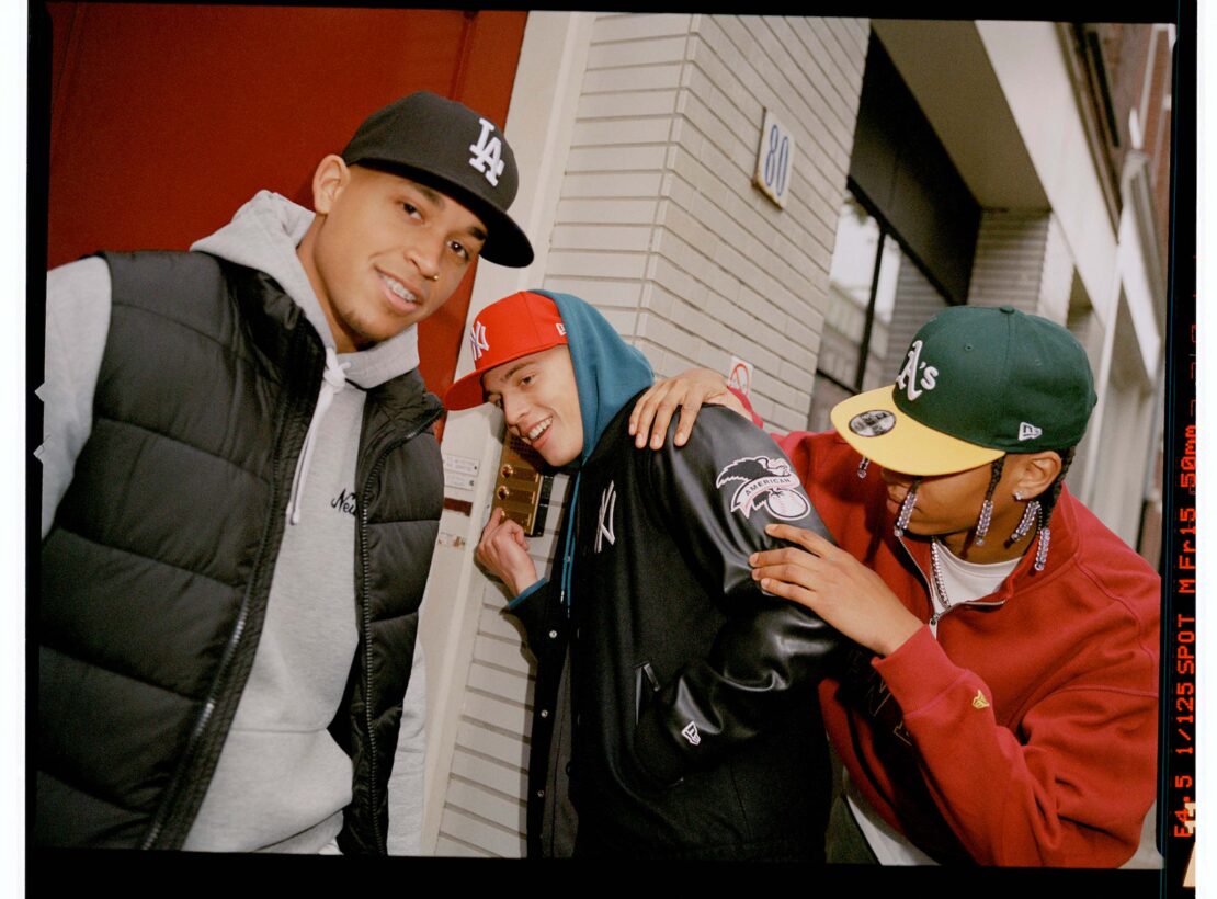 Three stylish young men pose in baseball caps and fashionable streetwear, standing outside near a building, showcasing urban fashion.