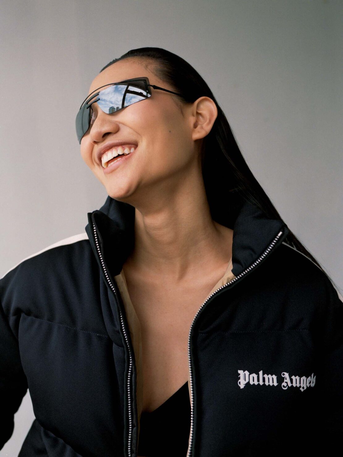 Smiling Asian woman with long black hair wears sunglasses and a black and white Palm Angels puffer jacket against a plain grey background.