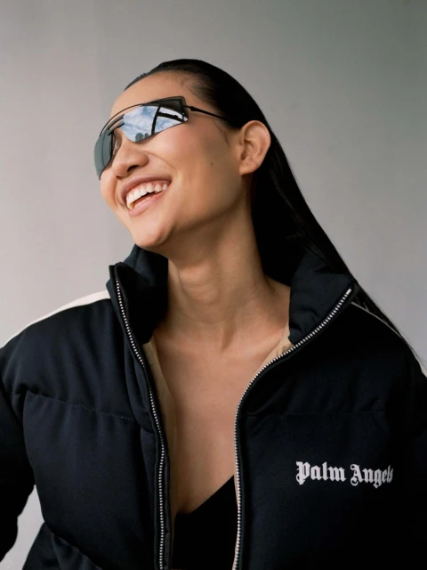 Smiling Asian woman with long black hair wears sunglasses and a black and white Palm Angels puffer jacket against a plain grey background.