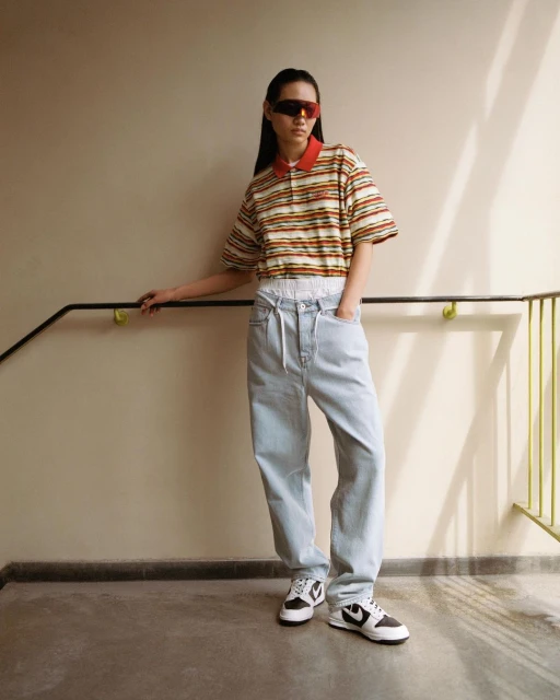 Woman in striped polo shirt, baggy jeans, and sunglasses poses against an indoor stairwell, showcasing a casual fashion style.