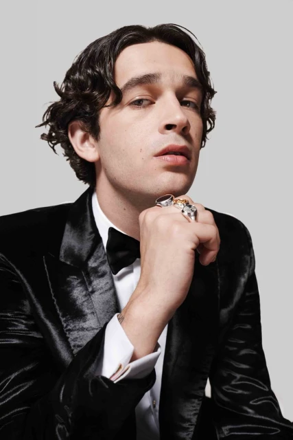 Portrait of a man in a black velvet tuxedo with a bow tie. He has dark curly hair, light skin, and is wearing several rings on his hand.