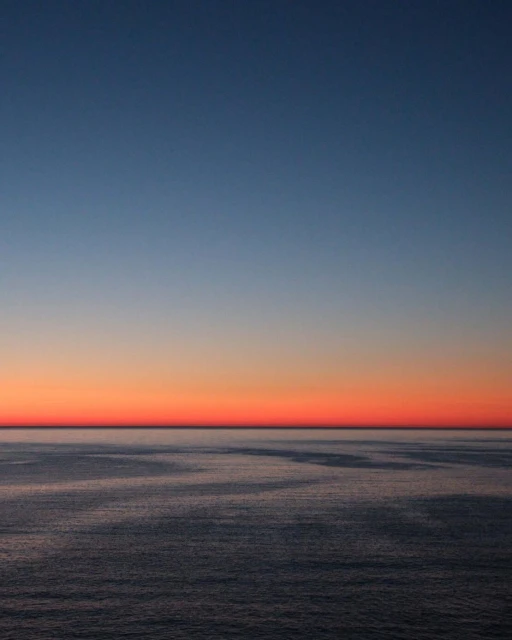 Scenic ocean view featuring the horizon with an orange and red sky fading into shades of blue during a vibrant sunset.