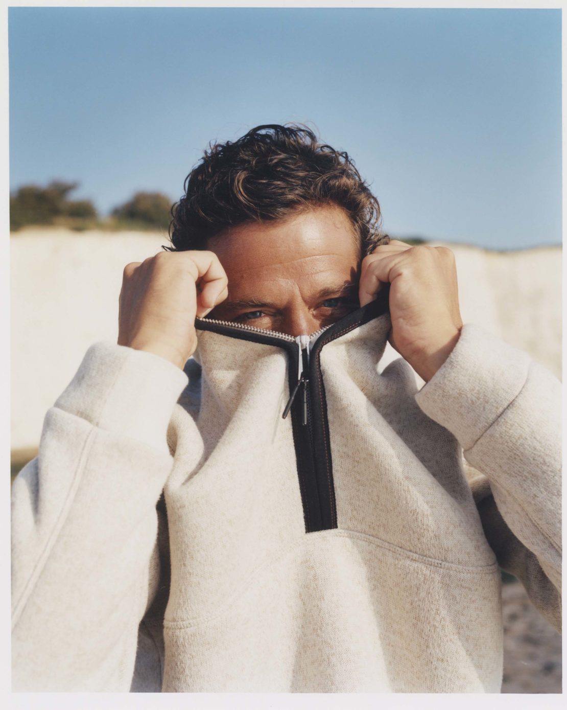 Man covering face with zippered sweatshirt against a pale blue sky and light-colored sand dune background. Outdoor portrait.